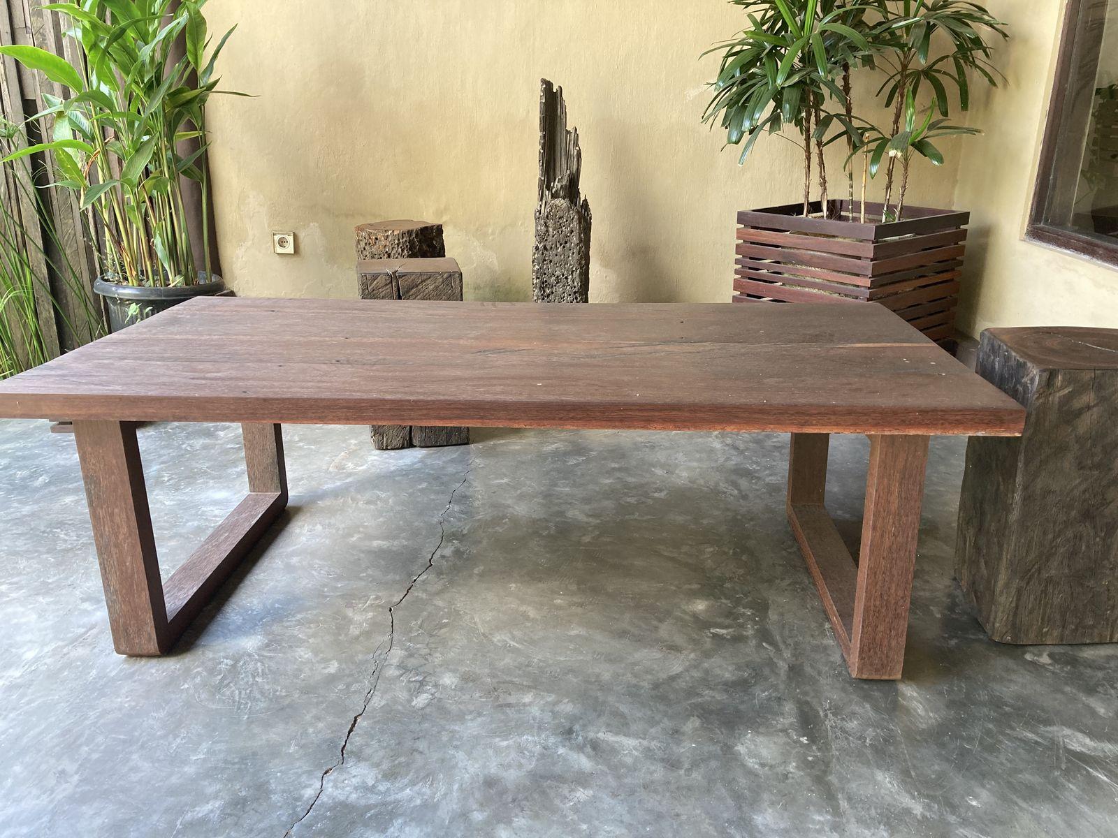 A finished dining table made from reclaimed ulin at Kaltimber in Bali, deep red-brown, standing on a polished concrete floor beside potted plants